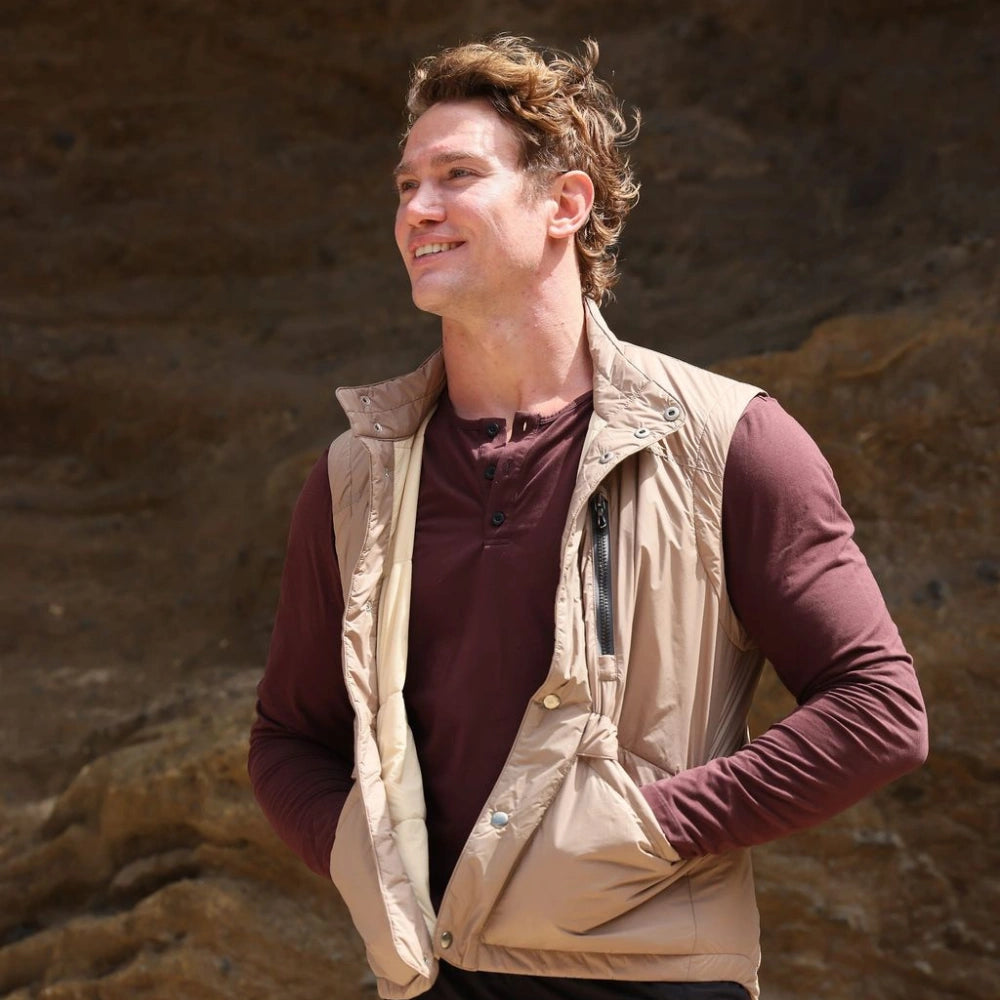 Man wearing a beige vest over a maroon shirt against a rocky background